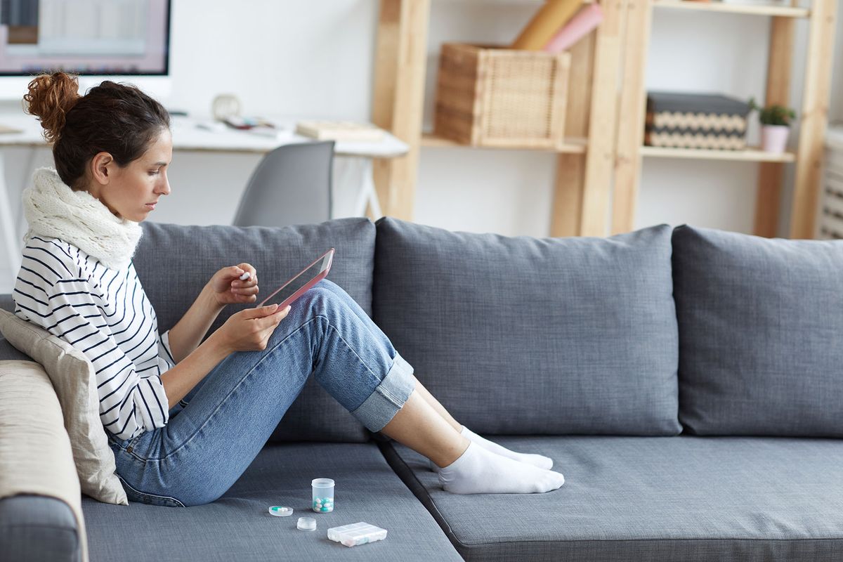 Ill young woman with pony tail wearing warm scarf around neck sitting on sofa with pill containers and browsing medical websites on tablet at home