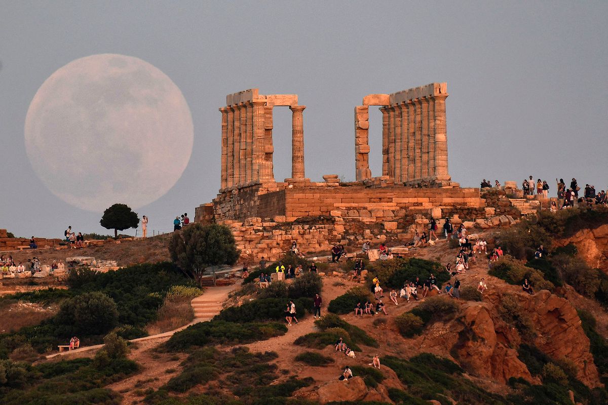 Locals and tourists watch the moon rising  by the Temple of Peseidon at the cape of Sounion, some 70 km south of Athens  on May 15, 2022. - A lunar eclipse is predicted on its set in early morning hours. (Photo by Louisa GOULIAMAKI / AFP)