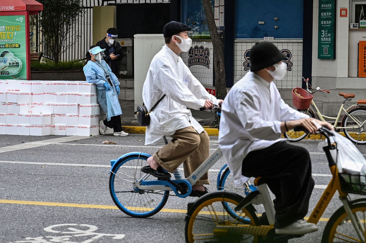 A worker wearing protective gear stands next to boxes to deliver in a neighborhood while two persons ride bicycle during a Covid-19 coronavirus lockdown in the Jing'an district in Shanghai on May 18, 2022. (Photo by Hector RETAMAL / AFP)