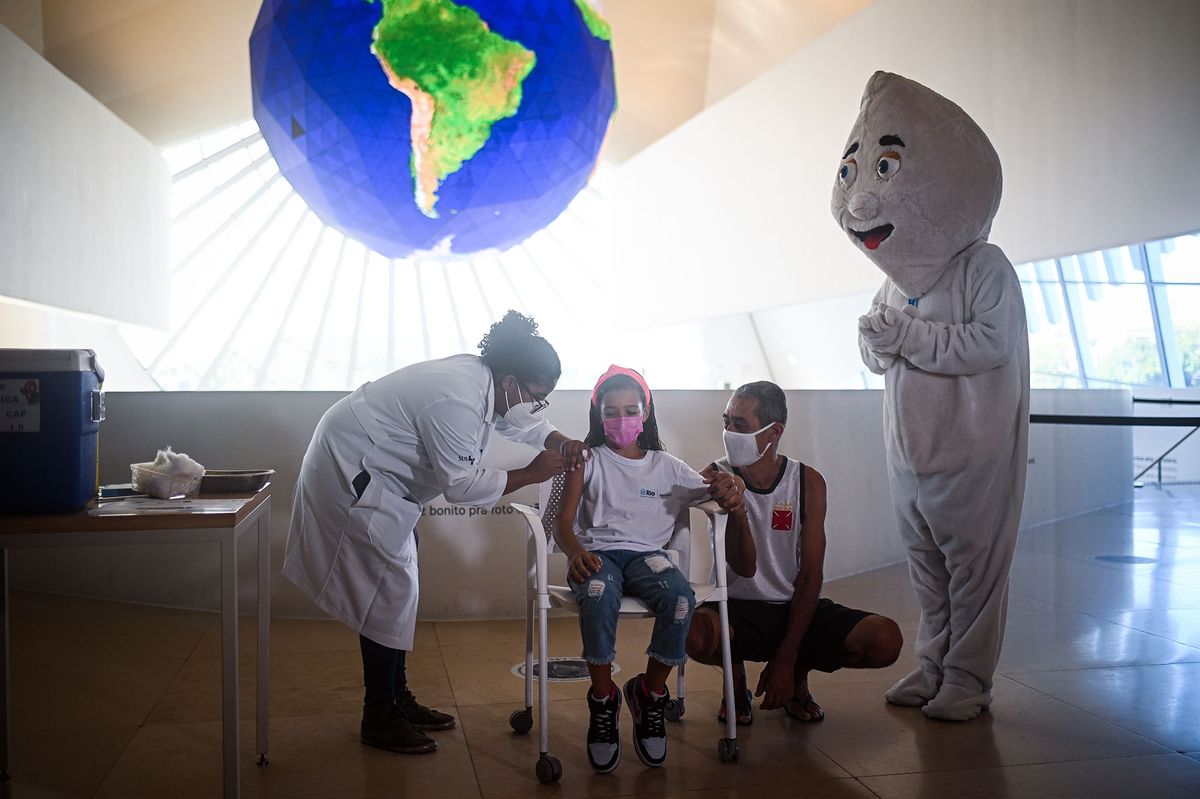 1237805628 A healthcare worker administers a dose of the Pfizer-BioNTech Covid-19 vaccine to a child beside her father and vaccination mascot Ze Gotinha at the Museu do Amanha (Museum of Tomorrow) in Rio de Janeiro, Brazil, on Monday, Jan. 17, 2022. Health Minister Marcelo Queiroga said Brazil will aim to have 20 million doses for children by the end March. Photographer: Andres Borges/Bloomberg via Getty Images