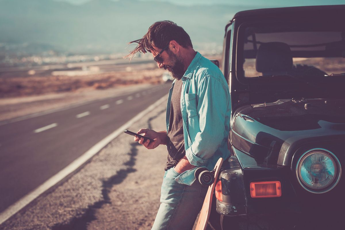 Adult man standing outdoor against black off road car using smartphone connection roaming technology to choose next travel destination. Concept of people and road trip vacation