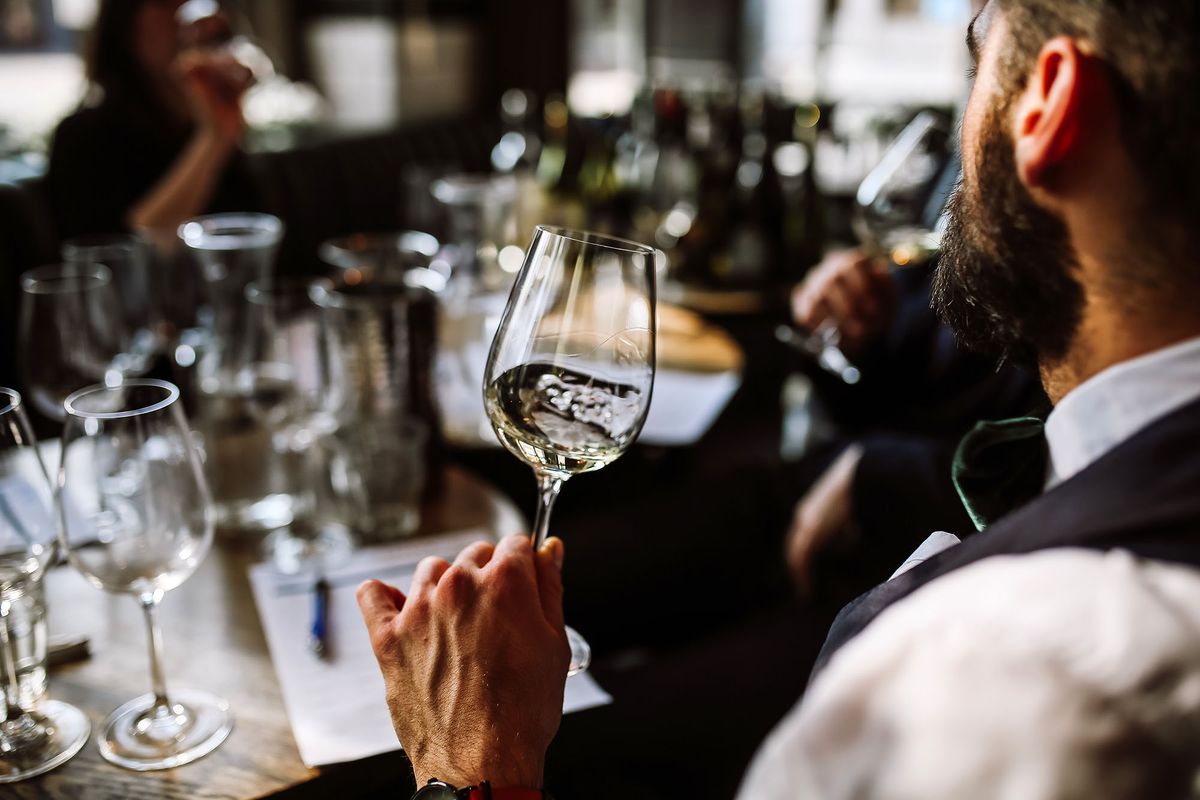 A young European man drinking white wine at the wine tasting. Selective focus point on the wine glass and the man. Other glasses and wine bottles in the background.
