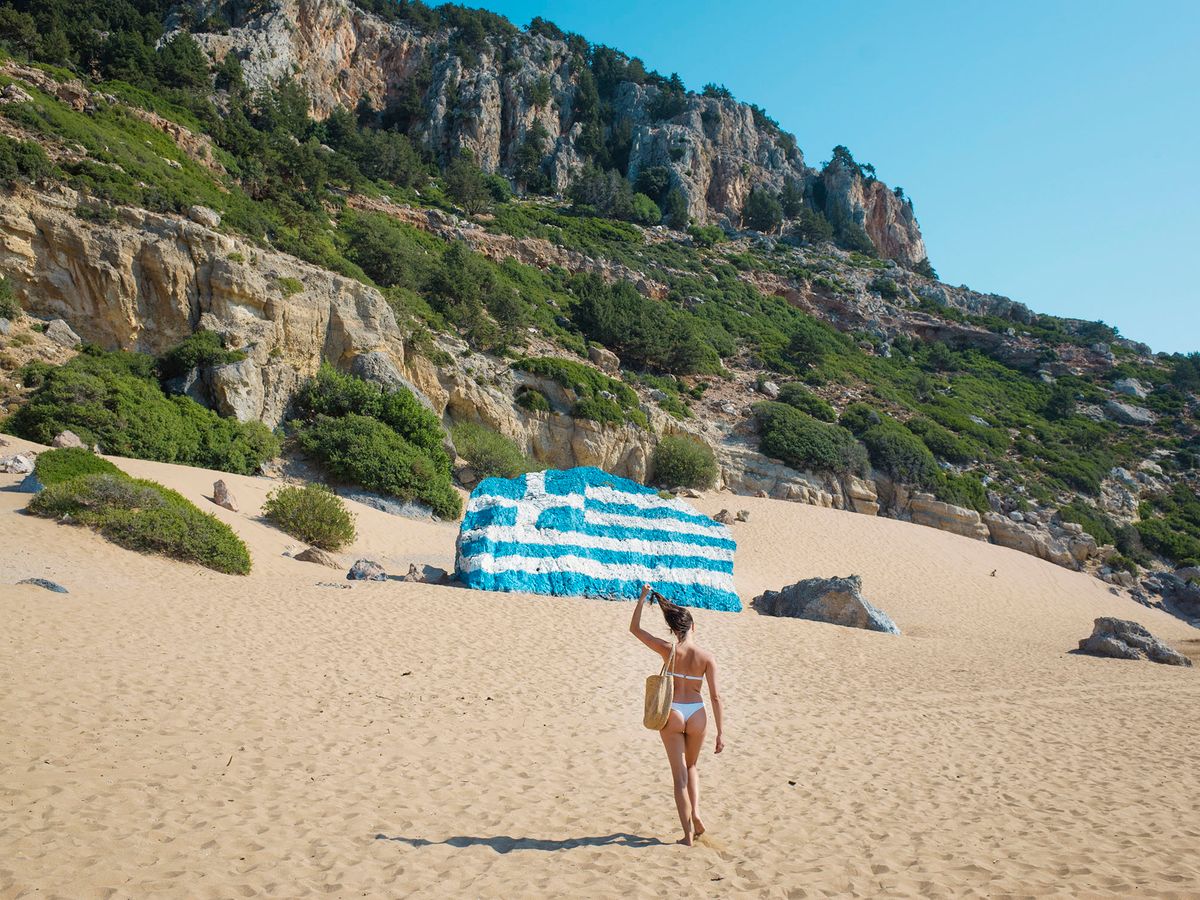 Rear view of young woman wearing bikini walking along sandy beach towards a boulder painted with the Greek flag. (Photo by Guido Cavallini / Cultura Creative / Cultura Creative via AFP)