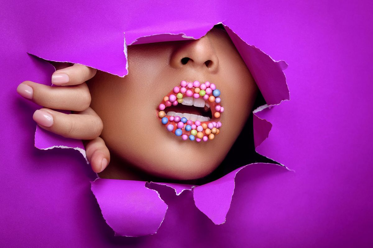 The sweet lips of the woman are covered with sweets. Creative makeup for advertising. Close-up of white teeth of an attractive woman through torn paper