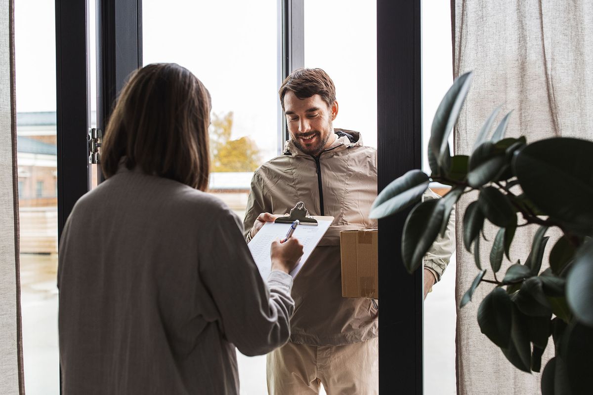 shipping, transportation and people concept - delivery man with parcel box and customer signing papers on clipboard at home
