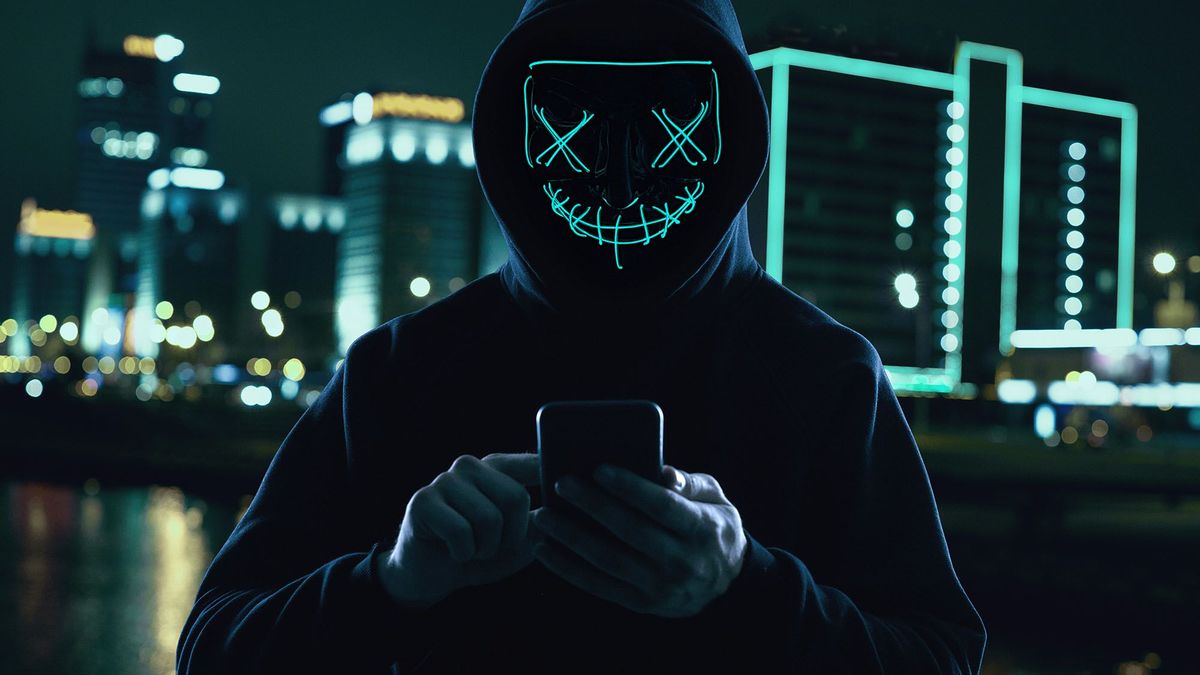 Portrait of an anonymous man in a black hoodie and neon mask hacking into a smartphone. Bright city background