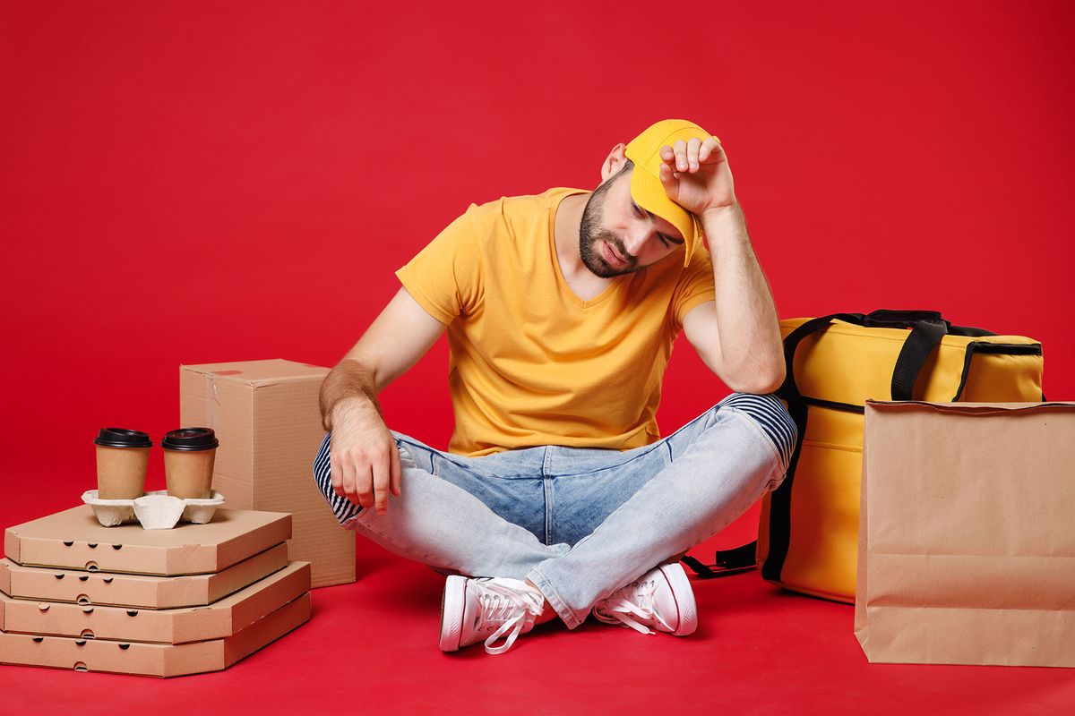 Delivery employee man guy male in yellow cap t-shirt sit at thermal takeaway food bag backpack pizza boxes cups work courier service during quarantine coronavirus covid-19 isolated on red background