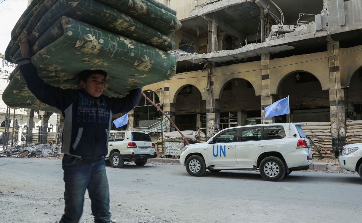 A Syrian boy carries mattresses over his head while walking past vehicles of the UN and the International Committee of the Red Cross (ICRC) delivering humanitarian aid in the Syrian town of Douma in the rebel-held enclave of Eastern Ghouta on the eastern outskirts of the capital Damascus on March 9, 2018. (Photo by Hamza AL-AJWEH / AFP)