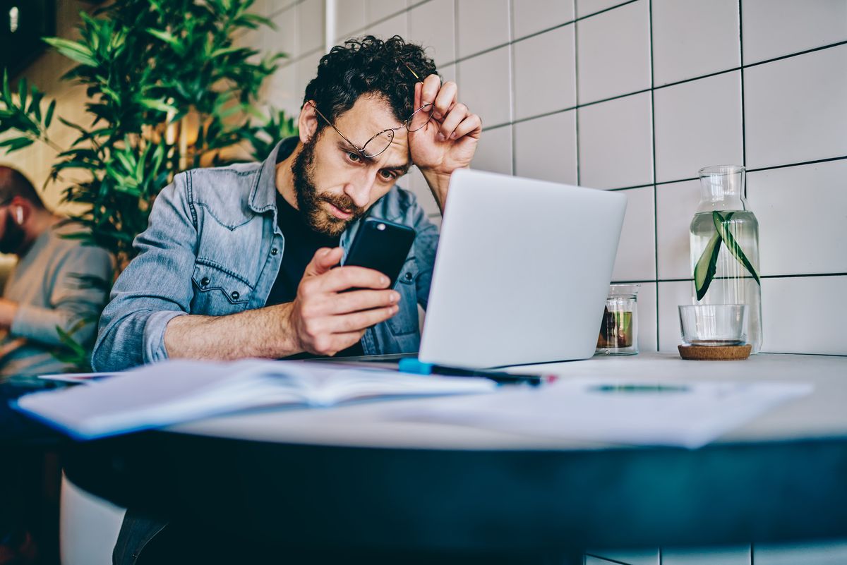 Upset,Young,Man,With,Eyeglasses,In,Hand,Reading,Business,News, Upset young man with eyeglasses in hand reading business news on website on modern mobile phone using free 4G internet sitting at laptop computer.Pensive hipster guy received warning message from bank