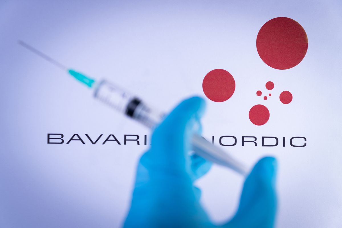 In this photo illustration a hand in medical gloves holds a syringe in front of Bavarian Nordic in Barcelona, Spain on September 2, 2021. Bavarian Nordic is a company developing a new Covid19 vaccine