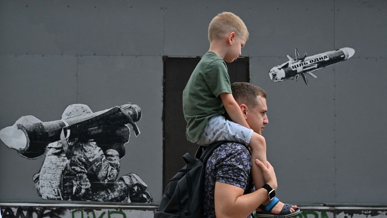 A man carries his son as they walk past a graffiti on a wall depicting a Ukrainian serviceman making a shot with a US-made Javelin portable anti-tank missile system, in Kyiv, on July 29, 2022. 