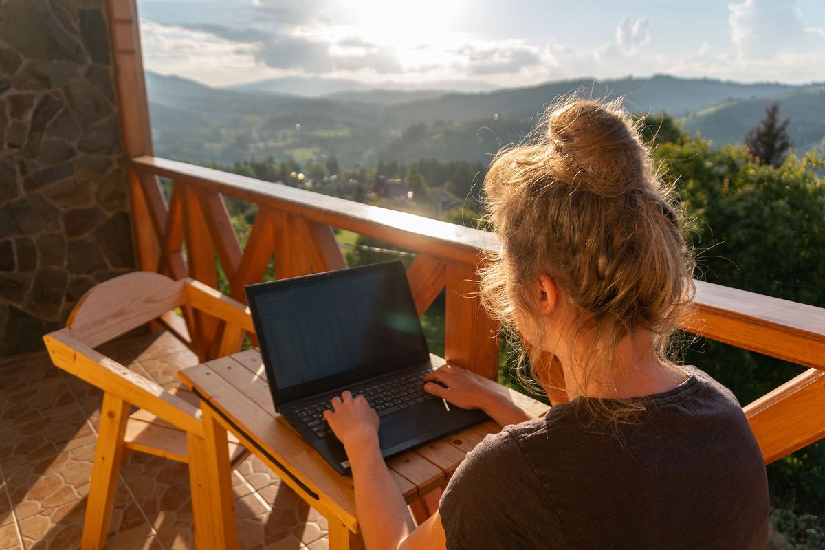 A young woman, freelancer is working on a laptop remotely on a balcony in the open fresh air near the mountains in warm sunny weather