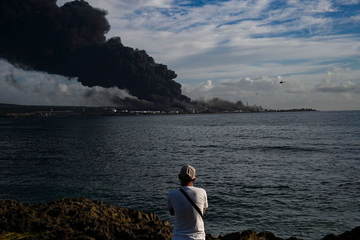 A man looks at black smoke rising from a massive fire at a fuel depot sparked by a lightning strike in Matanzas, Cuba, on August 8, 2022. - A second oil tanker collapsed at midnight Sunday in Matanzas, in western Cuba, where firefighters have been battling a huge blaze for two days, which has left one dead and 16 missing, local authorities reported. (Photo by YAMIL LAGE / AFP)