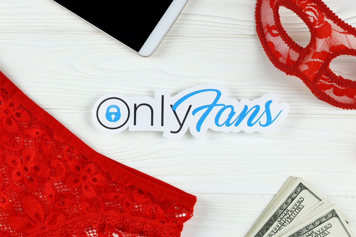 KHARKOV, UKRAINE - FEBRUARY 14, 2021: Onlyfans paper logo with dollar bills, red mask and red lingerie on white wooden table. OnlyFans is content subscription service based in London, UK