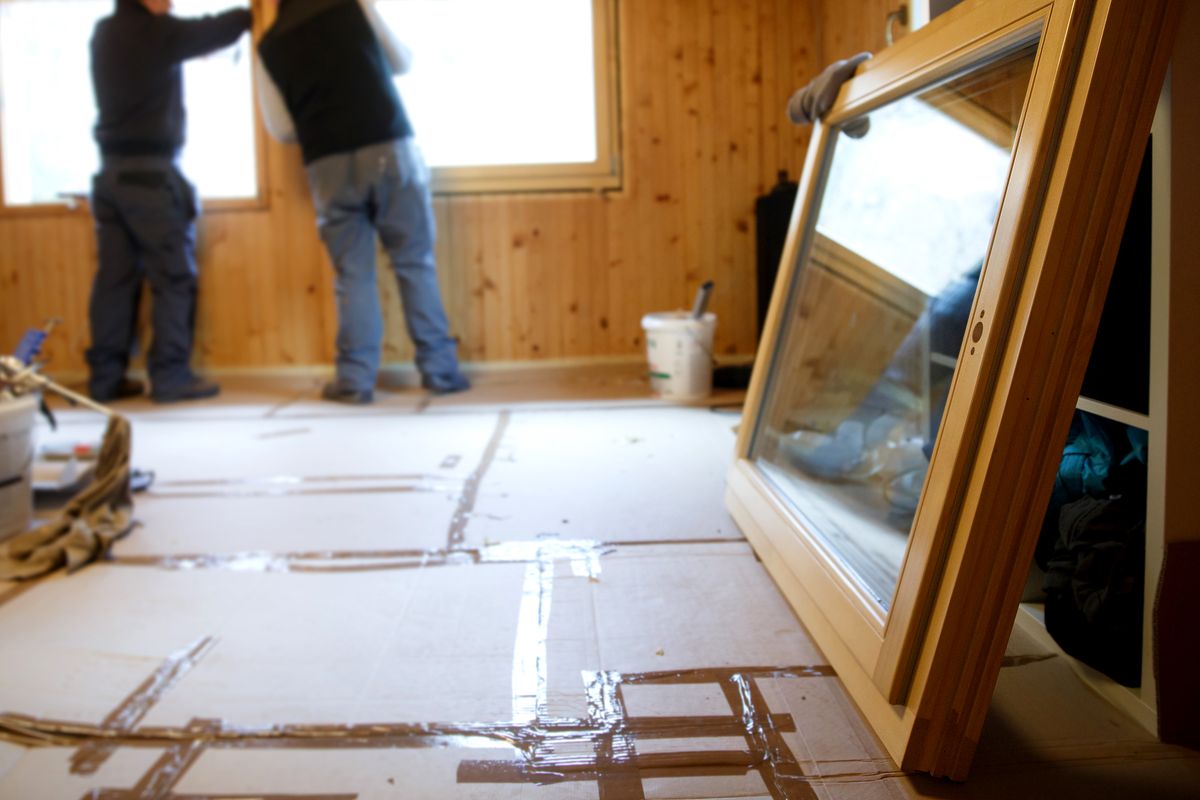 Workers in the background installing new, three pane wooden windows in an old wooden house, with a new window in the foreground. Home renovation, sustainable living, energy efficiency concept. ablakcsere, nyílászáró, ablak,