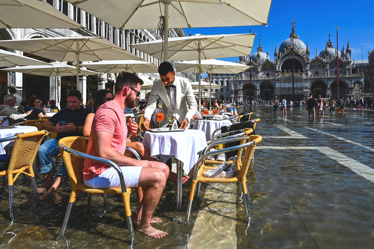 Tourists get their drinks served at a cafe's terrace on a flooded St. Mark's square in Venice on September 27, 2022, following an "Alta Acqua" high tide event, too low to operate the MOSE Experimental Electromechanical Module that protects the city of Venice from floods, but high enough to potentially threaten St. Mark's Basilica. (Photo by ANDREA PATTARO / AFP)
