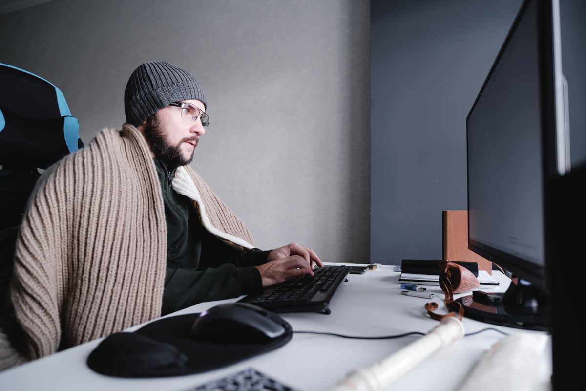 Young,Guy,In,Glasses,And,A,Warm,Hat,,Wrapped,In Young guy in glasses and a warm hat, wrapped in a blanket, works at a computer. It's cold in the room. The concept of turning off heating and gas, lowering the temperature in the room. Side view