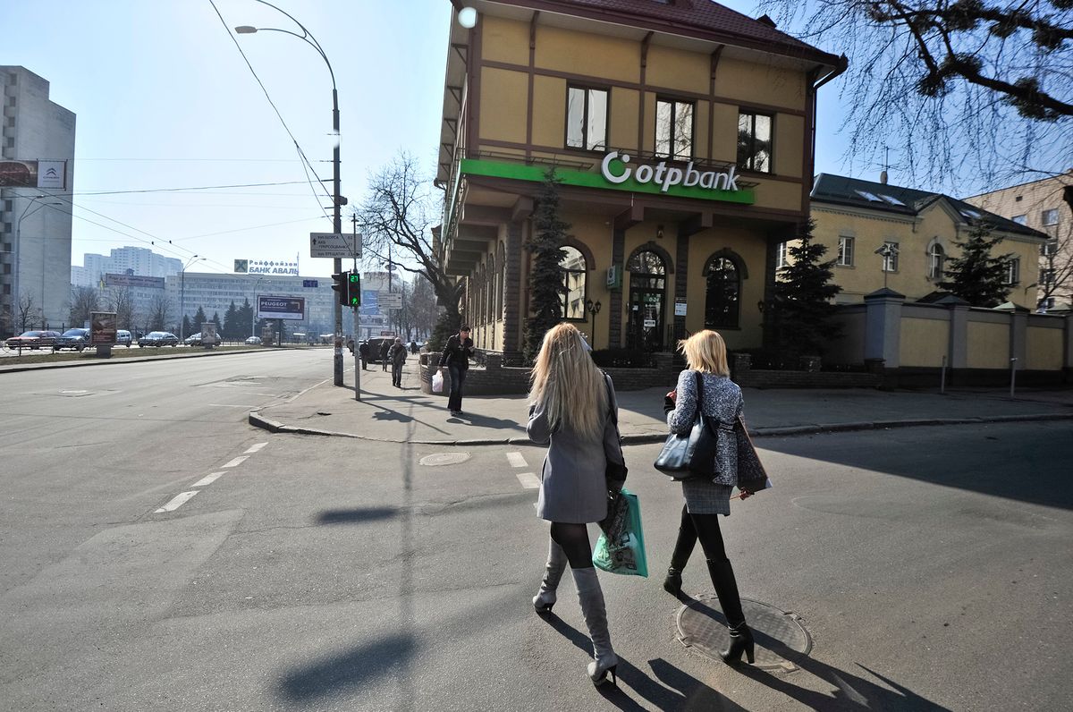Pedestrians cross a street outside an OTP Bank branch in Kiev, Ukraine, on Saturday, March 17, 2012. Ukraine said it wants to postpone $3 billion of bailout repayments to the International Monetary Fund, as Standard & Poor's cut its outlook on the former Soviet State's debt to negative, citing "significant" funding risks. Photographer: 
