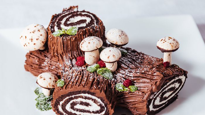 French,Dessert,Called,Yule,Log,Or,Bűche,De,Noël,With French dessert called Yule log or bûche de Noël with merengue mushrooms and mint leaves on top of chocolate glazing. Placed in front of Christmas tree. Decorated for Christmas Holidays or New year