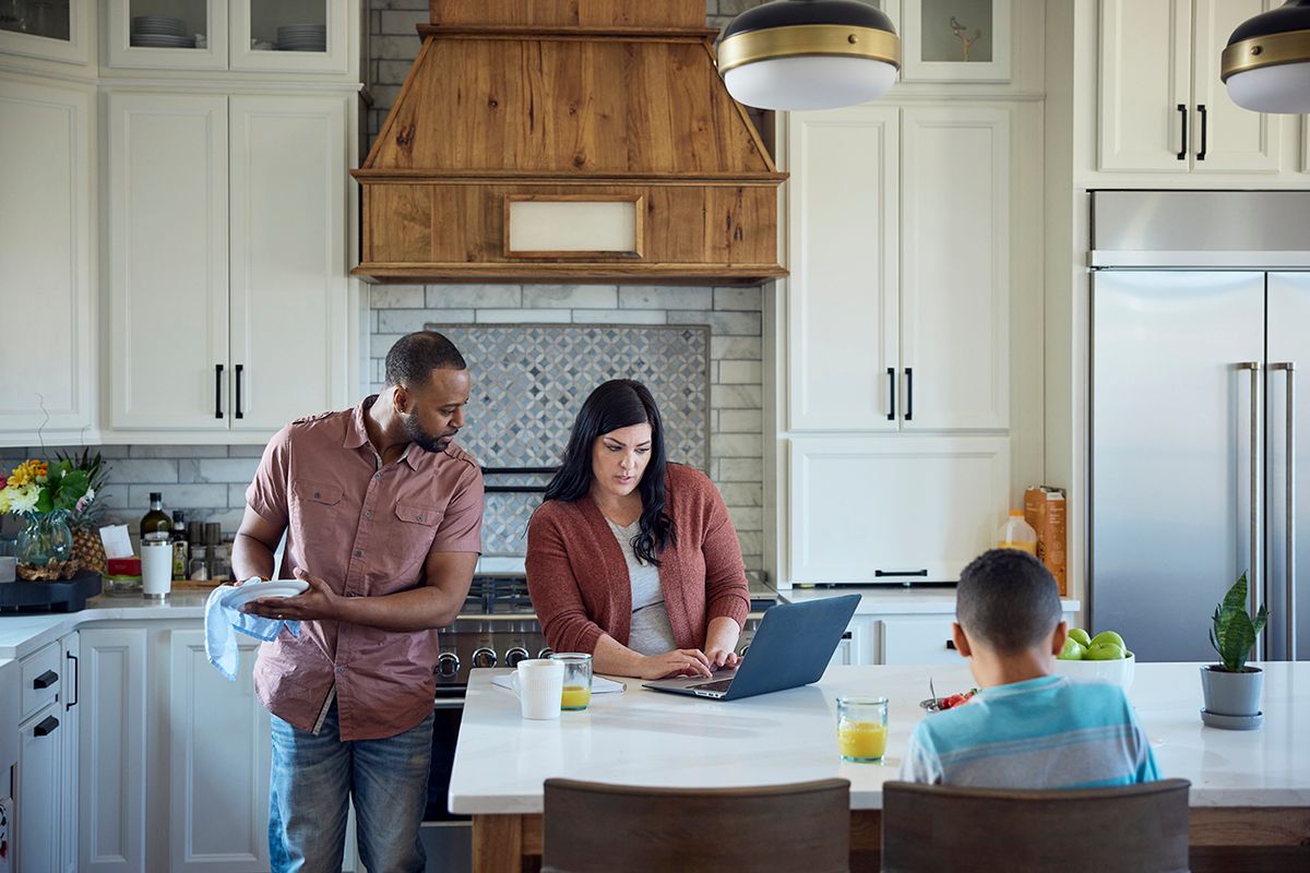 African American dad washes dishes while Caucasian mom works on laptop and young son eats breakfast. African American dad washes dishes while Caucasian mom works on laptop and young son eats breakfast.