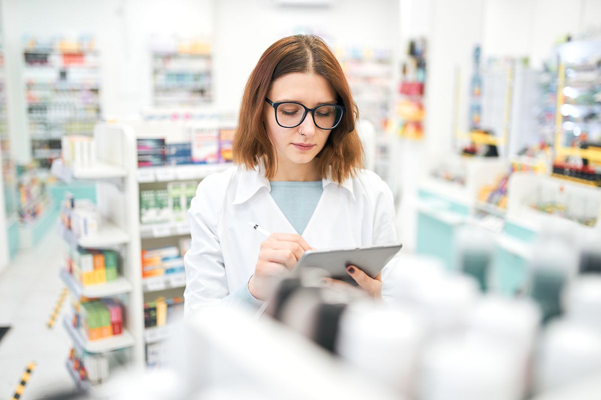 Young pharmacist browsing data on tablet during work Focused young woman in uniform and glasses making notes and browsing data on tablet while inventorying medical supplies in drugstore.