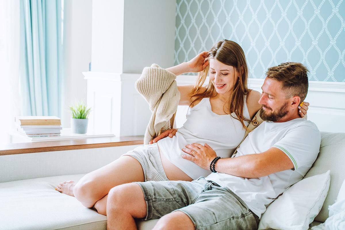 Happy family of man and pregnant woman sitting on a sofa at home in light modern room Bearded man touching belly of pregnant woman with love. Romantic couple in relationship waiting for a baby