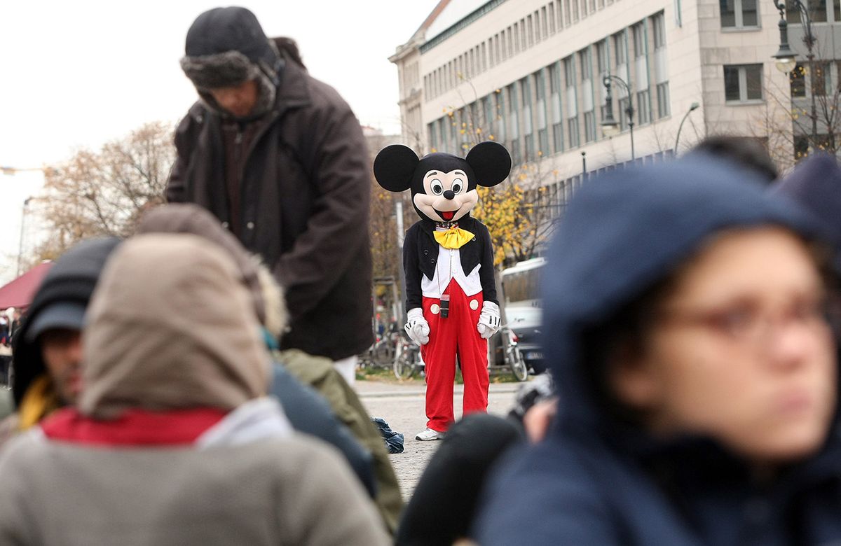 Protesting Asylum Seekers Launch Hunger Strike BERLIN, GERMANY - OCTOBER 25: A street performer dressed as Mickey Mouse stands near protesting refugees as they stage a hunger strike in front of the Brandenburg Gate on October 25, 2012 in Berlin, Germany. The demonstrators, predominantly from Iran, Afghanistan and Iraq are subsisting on only water, tea and coffee without sugar, and claim to have been told by the police that they are not allowed blankets or tents. They have been sitting in front of the Brandenburg Gate since last night, and say they will continue their strike until the German government responds to their demands for a halt to deportations and faster processing of asylum applications.  (Photo by Adam Berry/Getty Images)