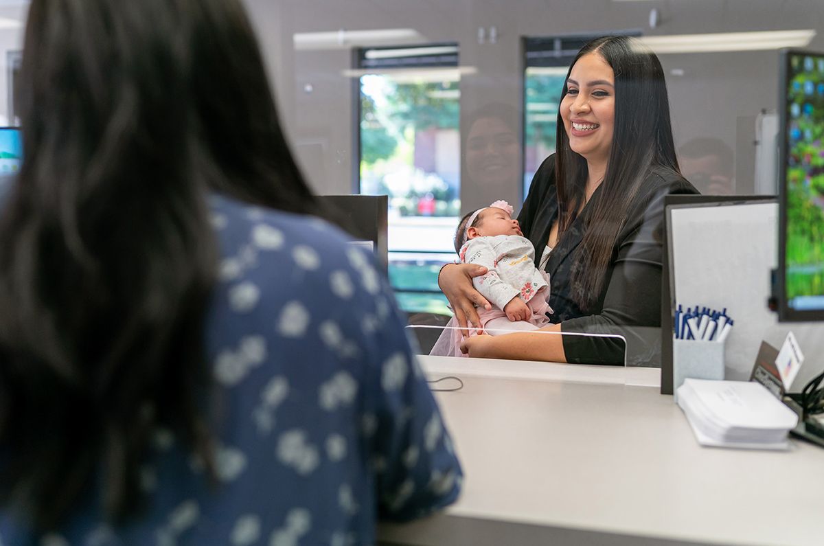 Hispanic woman with baby at business reception desk A beautiful young woman of Hispanic descent holds her baby and smiles while speaking to a customer service representative at a medical office or bank reception desk outfitted with plexiglass sneeze guard for safety.