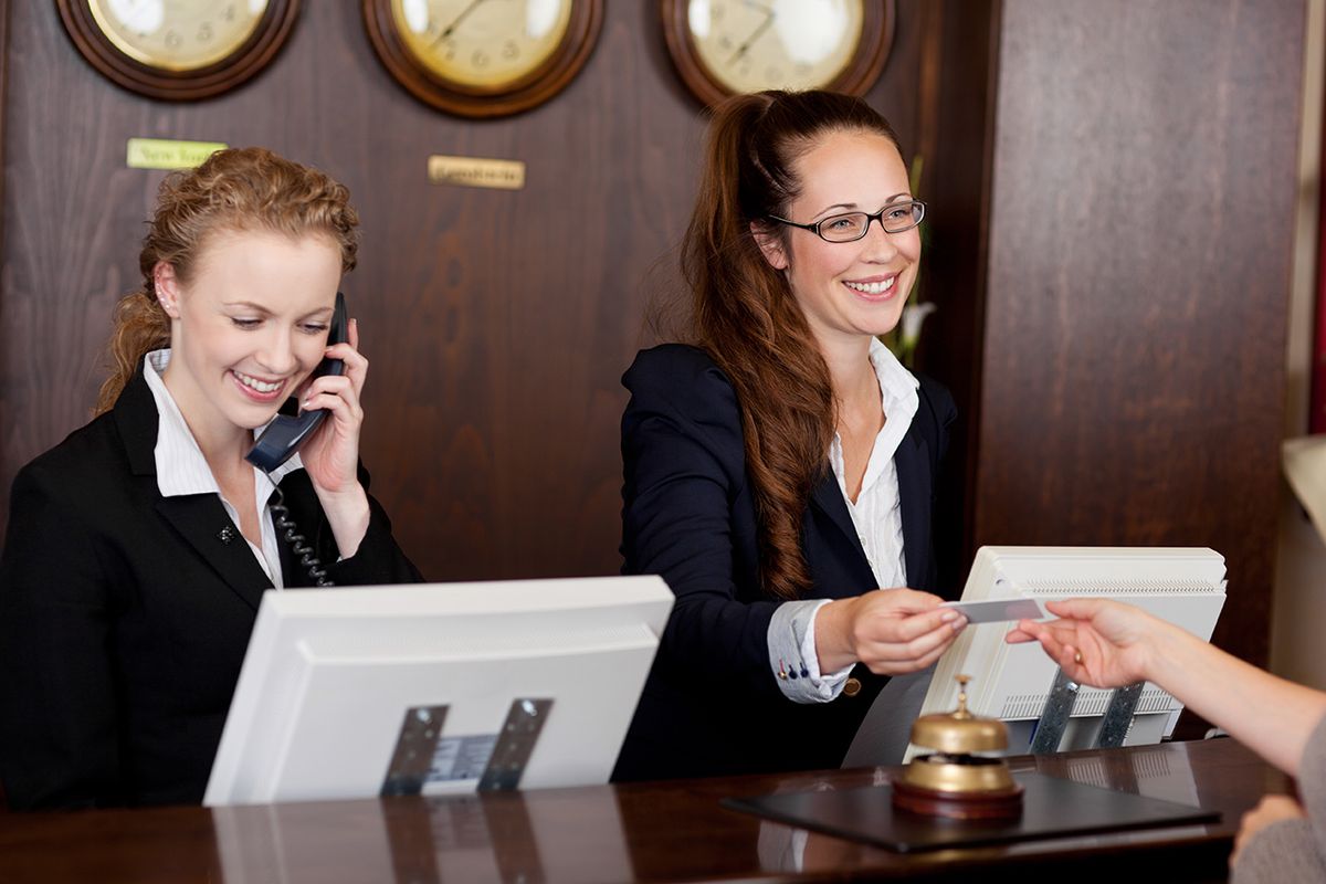 Two,Beautiful,Young,Stylish,Receptionists,At,A,Reception,Desk,,One Two beautiful young stylish receptionists at a reception desk, one talking on the telephone and the other handing a card to a customer
