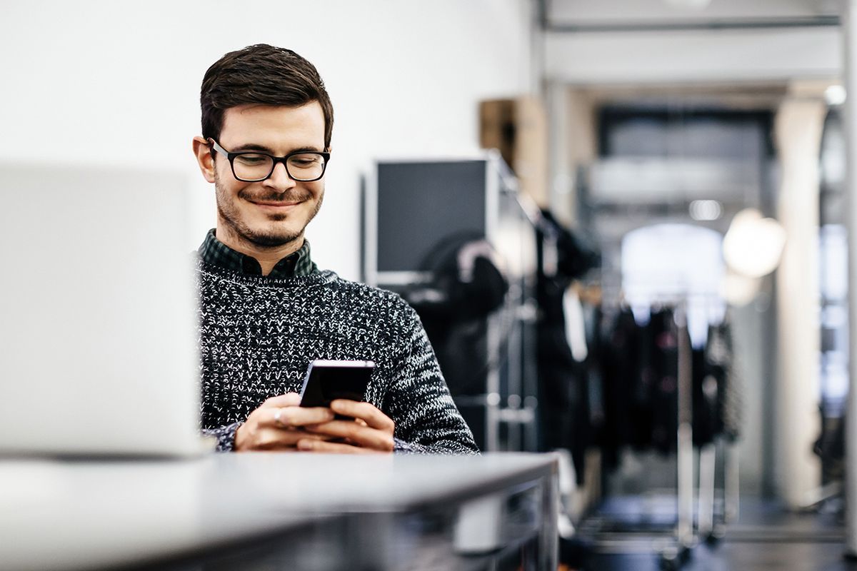 Young casual businessman looking at his smartphone A young casual businessman wearing glasses is smiling at his smartphone in a bright modern office room. Clothes racks are seen in the background and a notebook in the front.