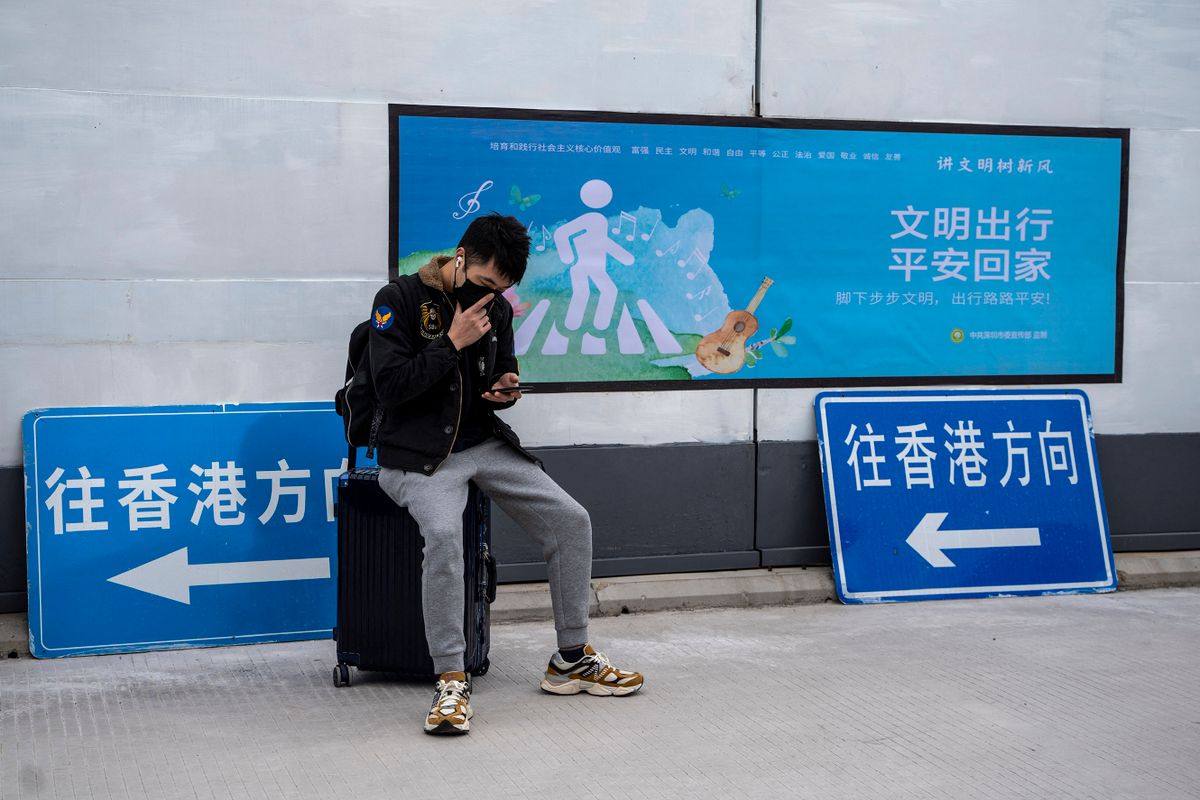 A man sitting on a suitcase next to signs pointing to Hong Kong at Shenzhen Bay Port on January 8, 2023 in Shenzhen, China. Hong Kong today resumes quarantine free travel with China, but still requiring travelers to produce a Negative PCR Covid Test Result and register for a crossing quote. 