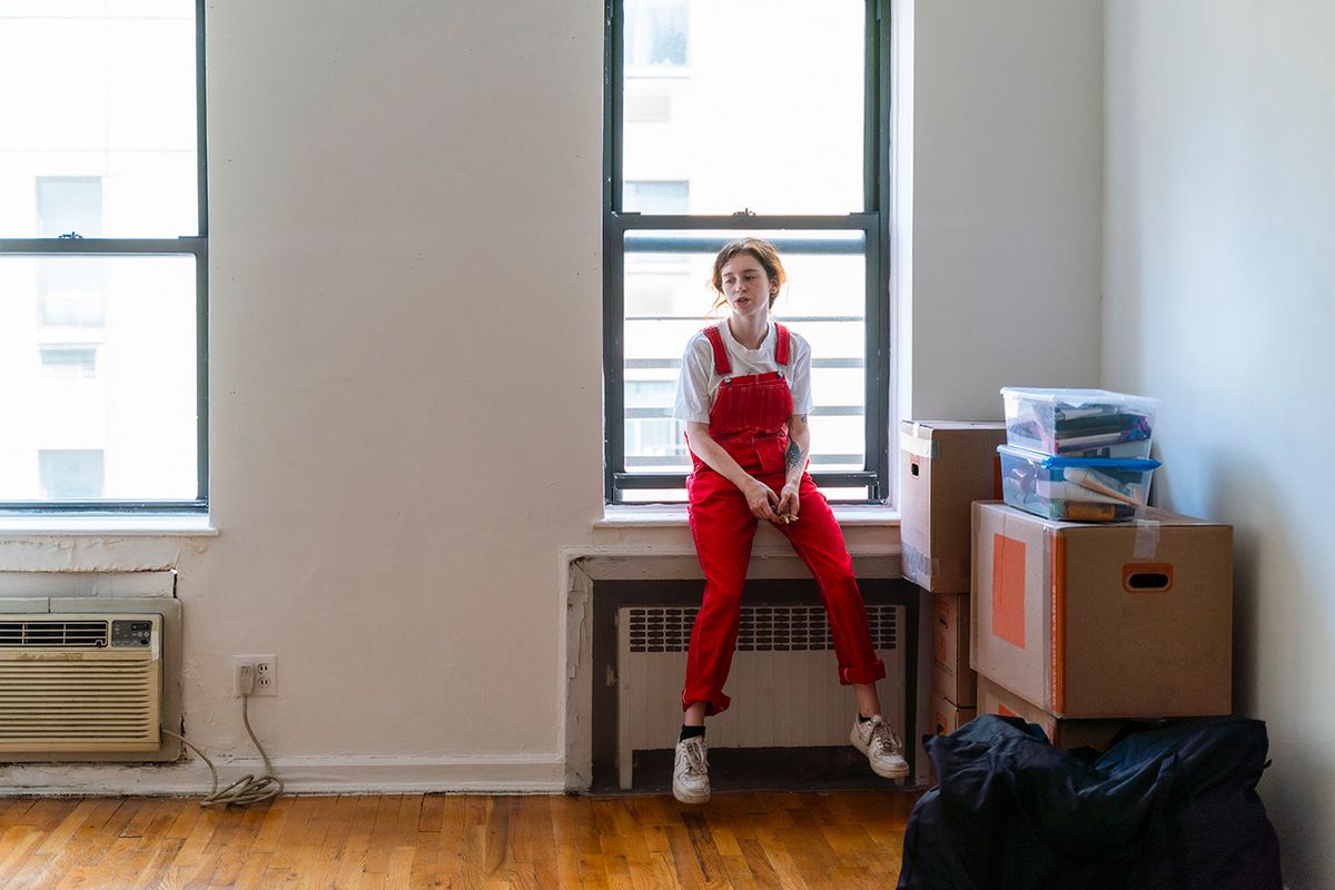Relocation to a new apartment. Young woman is sitting on a windowsill in an empty room of a newly rented apartment, tired after moving in. Young woman in a red bib overall is sitting on a windowsill next to Items packed to move are on the floor. She is taking a rest.