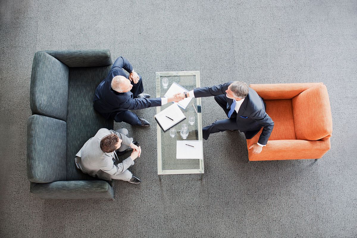 Businessmen shaking hands across coffee table in office lobby
vállalatfelvásárlás, üzletkötés