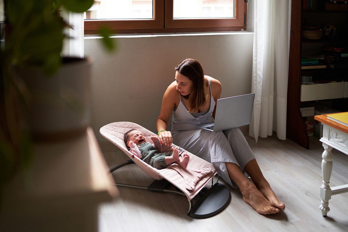 Mother using a laptop next to a baby lying on a baby swing at home Mother using a laptop while consoling a baby lying on a baby swing at home