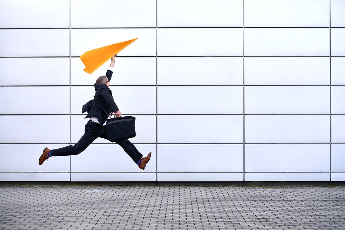 Businessman in black suit running fast and jumping with a paper  plane and a laptop bag Businessman running fast and jumping up with an orange paper plane and a laptop bag. Funny business concept.
