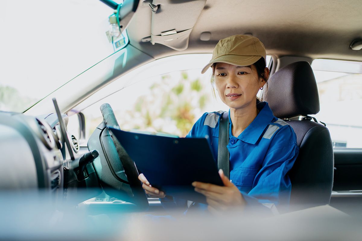 A female courier in blue uniform and baseball cap checking on customer delivery address while seating on the driver’s seat A female delivery woman wearing baseball cap checking on customer delivery address while seating on the driver’s seat before making a home delivery.