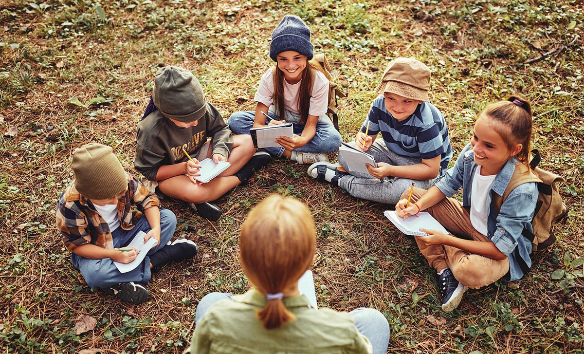 School,Activities,In,Nature.,Group,Of,Curious,Children,And,Young School activities in nature. Group of curious children and young female teacher sitting together on green grass in forest and learning about environment, boys and girls during ecology lesson in wood