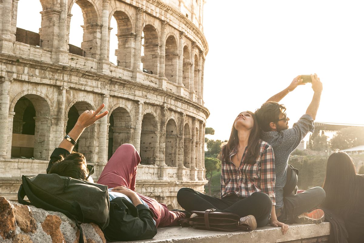 Three,Young,Friends,Tourists,Sitting,Lying,In,Front,Of,Colosseum
Three young friends tourists sitting lying in front of colosseum in rome taking selfie pictures with smartphone camera. Sunset with lens flare.