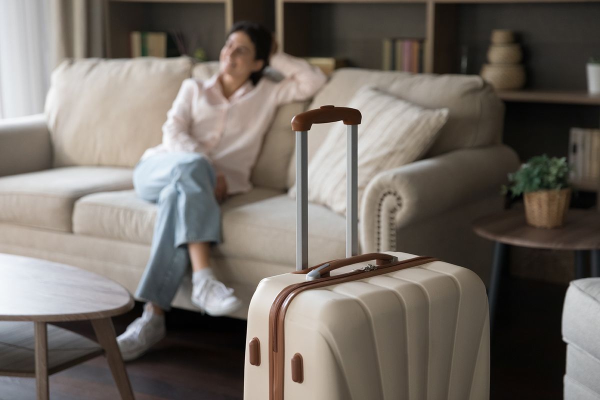 Focus,On,Beige,Suitcase,With,Blurred,Carefree,Young,Woman,Sitting Focus on beige suitcase with blurred carefree young woman sitting on sofa on background. Joyful refreshed millennial Hispanic lady satisfied with vacation travel, relaxing alone in modern hotel room.