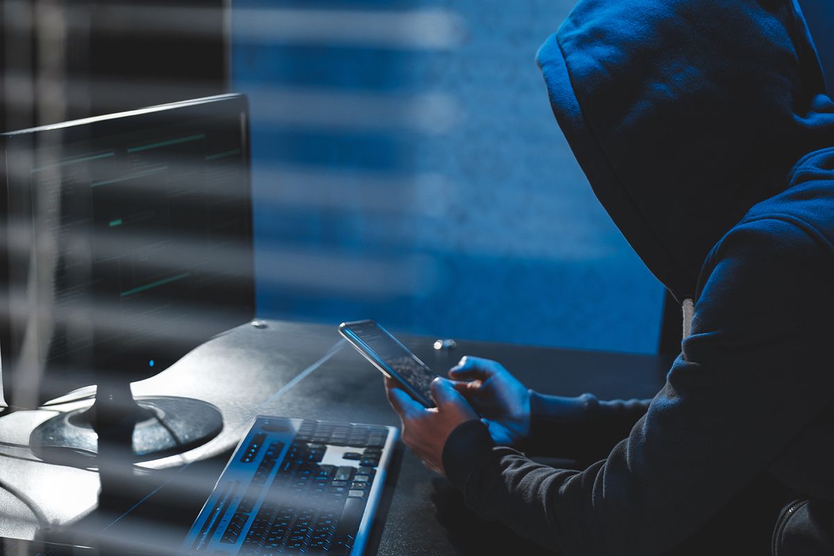 Overhead hacker in hood working at computer and mobile phone typing text in dark room, An anonymous hacker uses malware with mobile phone to hack password, personal data steals money from bank. cyber