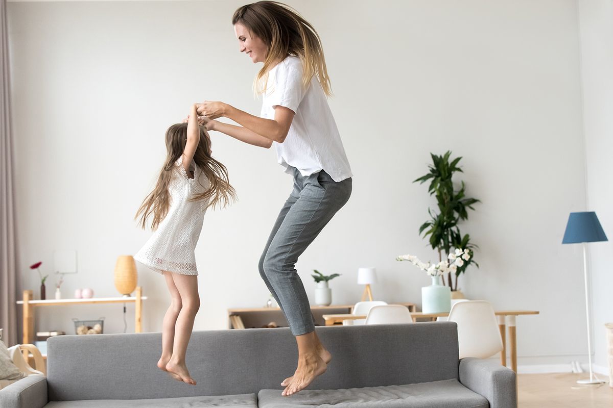 Happy mommy and kid daughter holding hands jumping on sofa together, baby sitter or mother playing having fun with cute kid girl at home, young mum and child enjoy spending time laughing together
