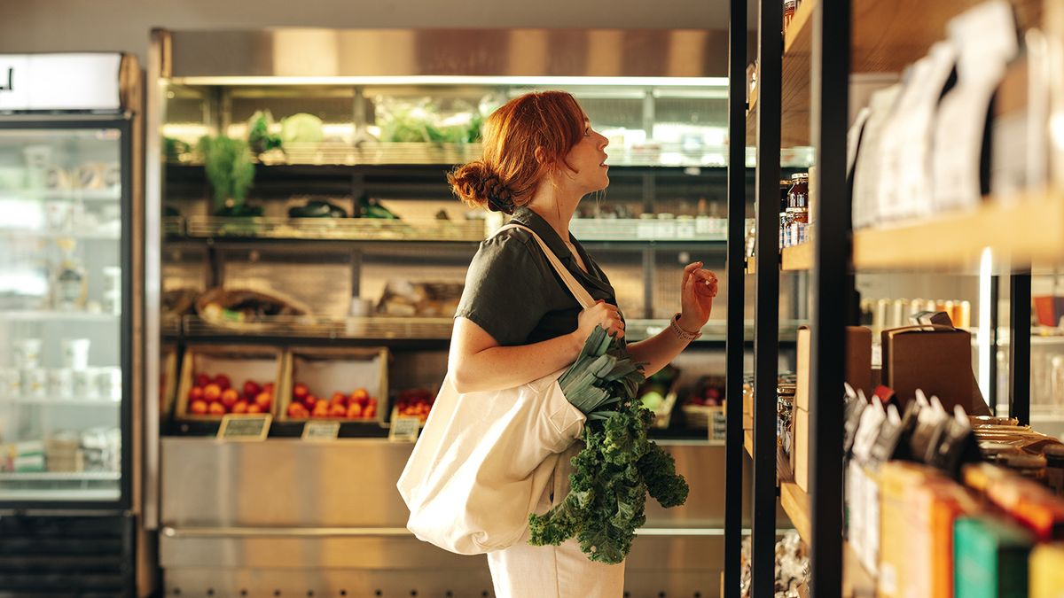 Female,Shopper,Choosing,Food,Products,From,A,Shelf,While,Carrying
Female shopper choosing food products from a shelf while carrying a bag with vegetables in a grocery store. Young woman doing some grocery shopping in a trendy supermarket.
