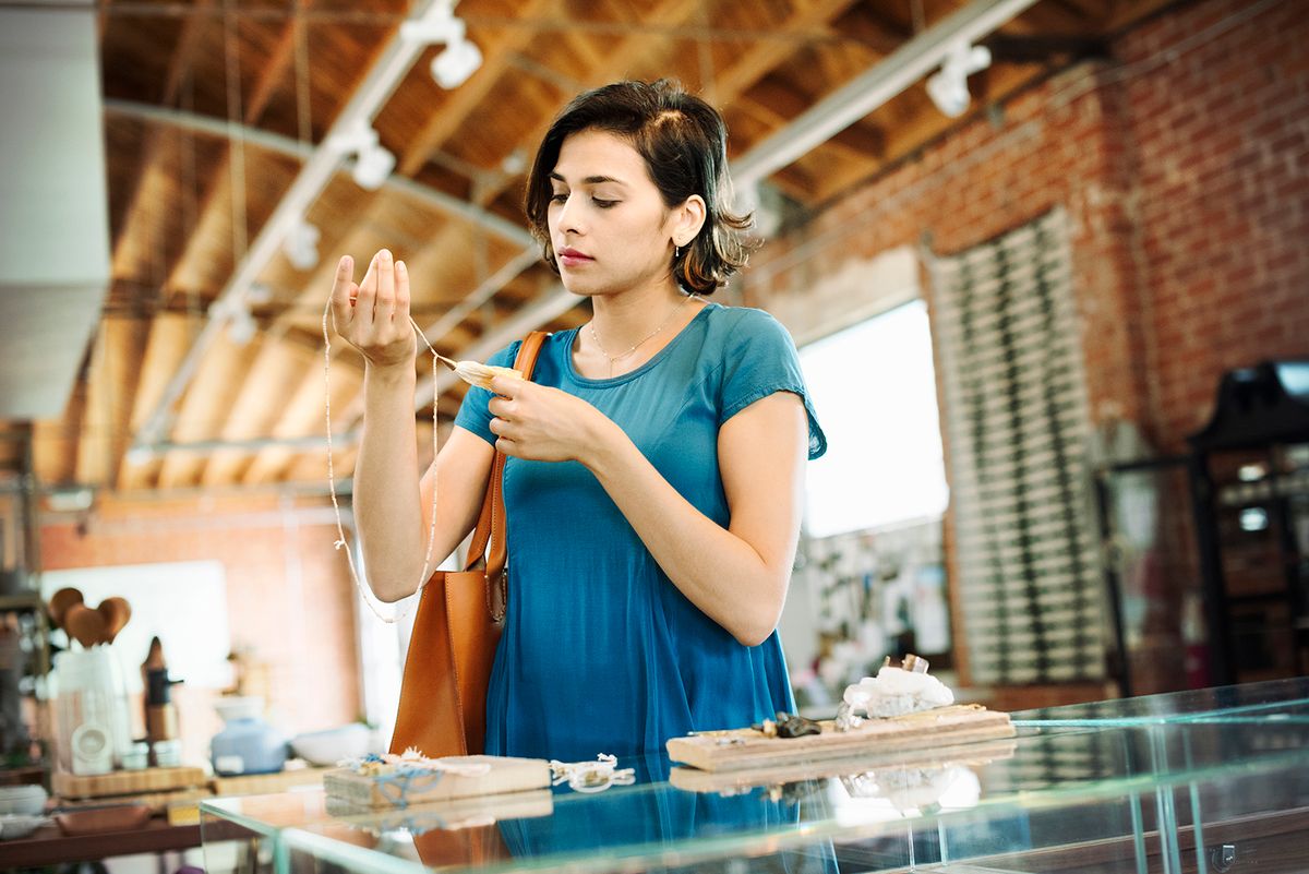 Young woman standing in a shop, looking at a necklace.Young woman standing in a shop, looking at a necklace.
Mint Images,GTTY