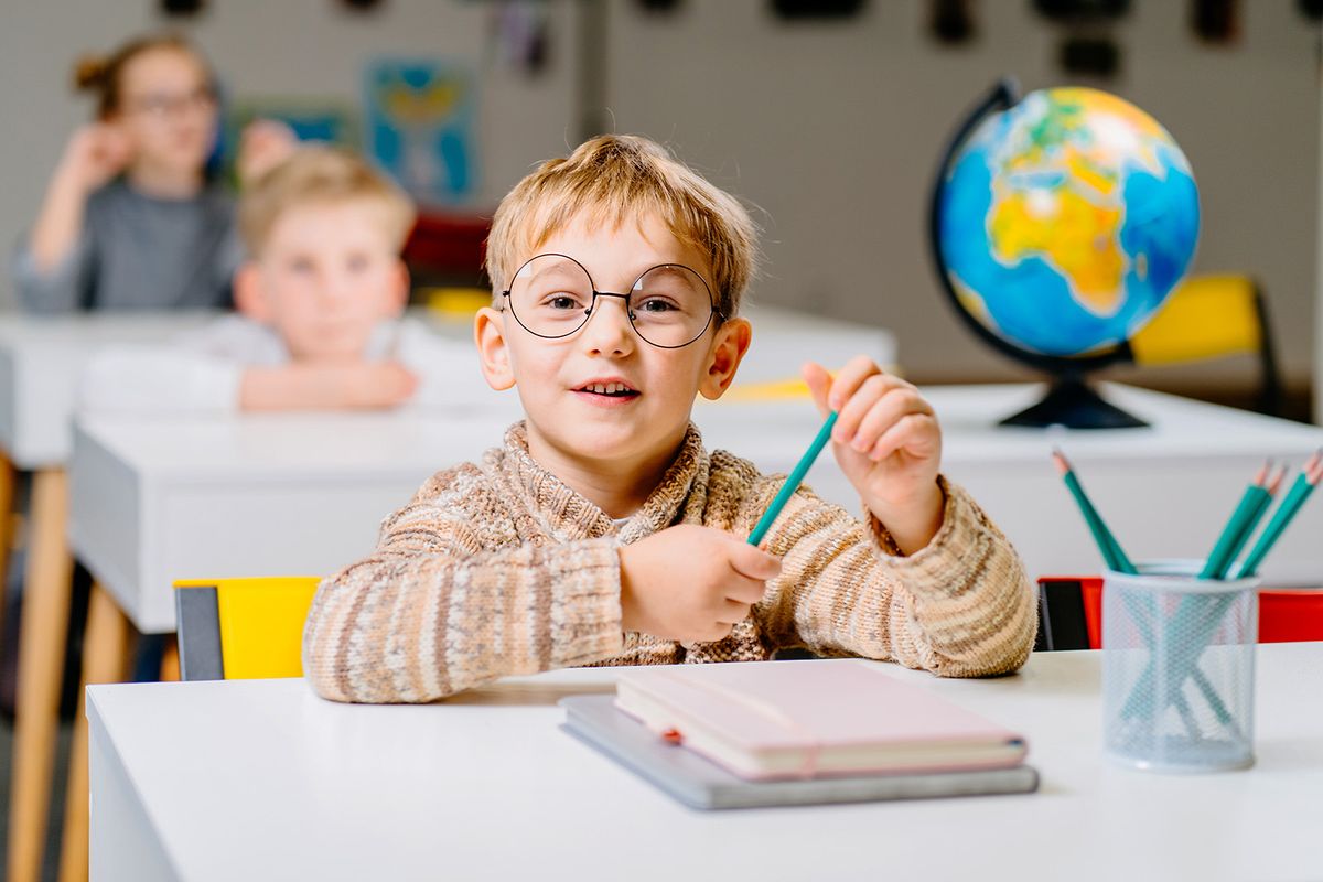 Cute blond male student in eyeglasses in class with globe in elementary school. Portrait of little schoolboy studying with classmates in background.