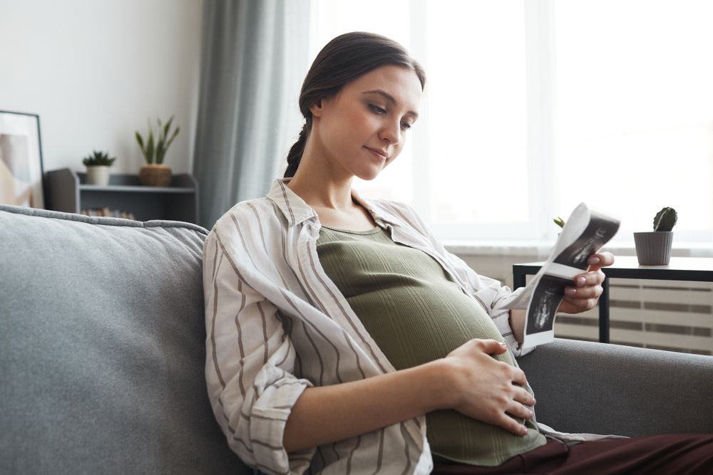 Pregnant,Woman,Sitting,On,Sofa,And,Looking,At,Ultrasound,Image