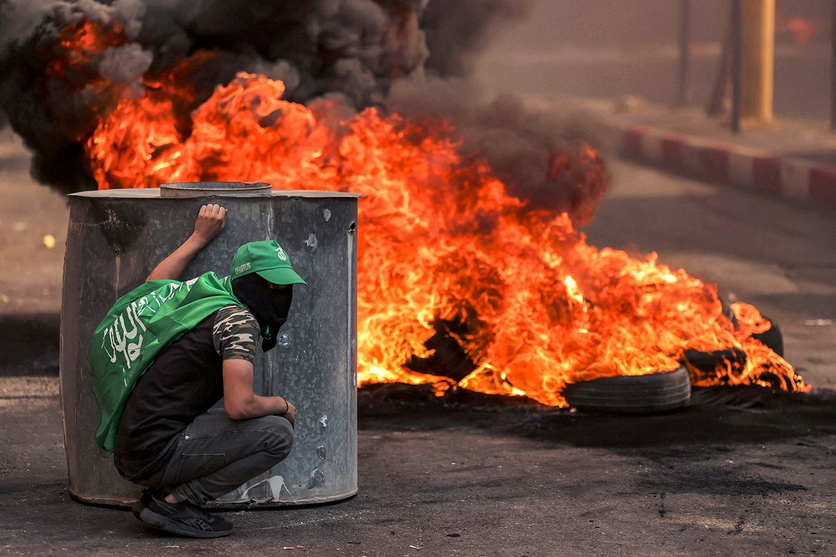 A masked Palestinian protester takes cover near flaming tires during clashes with Israeli forces following a rally in solidarity with Gaza by supporters of the Fatah and Hamas movements, in the city of Hebron in the occupied West Bank on October 13, 2023. AFP correspondents and a security official reported clashes after rallies in solidarity with war-battered Gaza in Ramallah, Nablus, Tulkarm, Hebron and other cities and towns, with the Palestinian Red Crescent reporting dozens wounded across the West Bank, some critically. (Photo by HAZEM BADER / AFP)