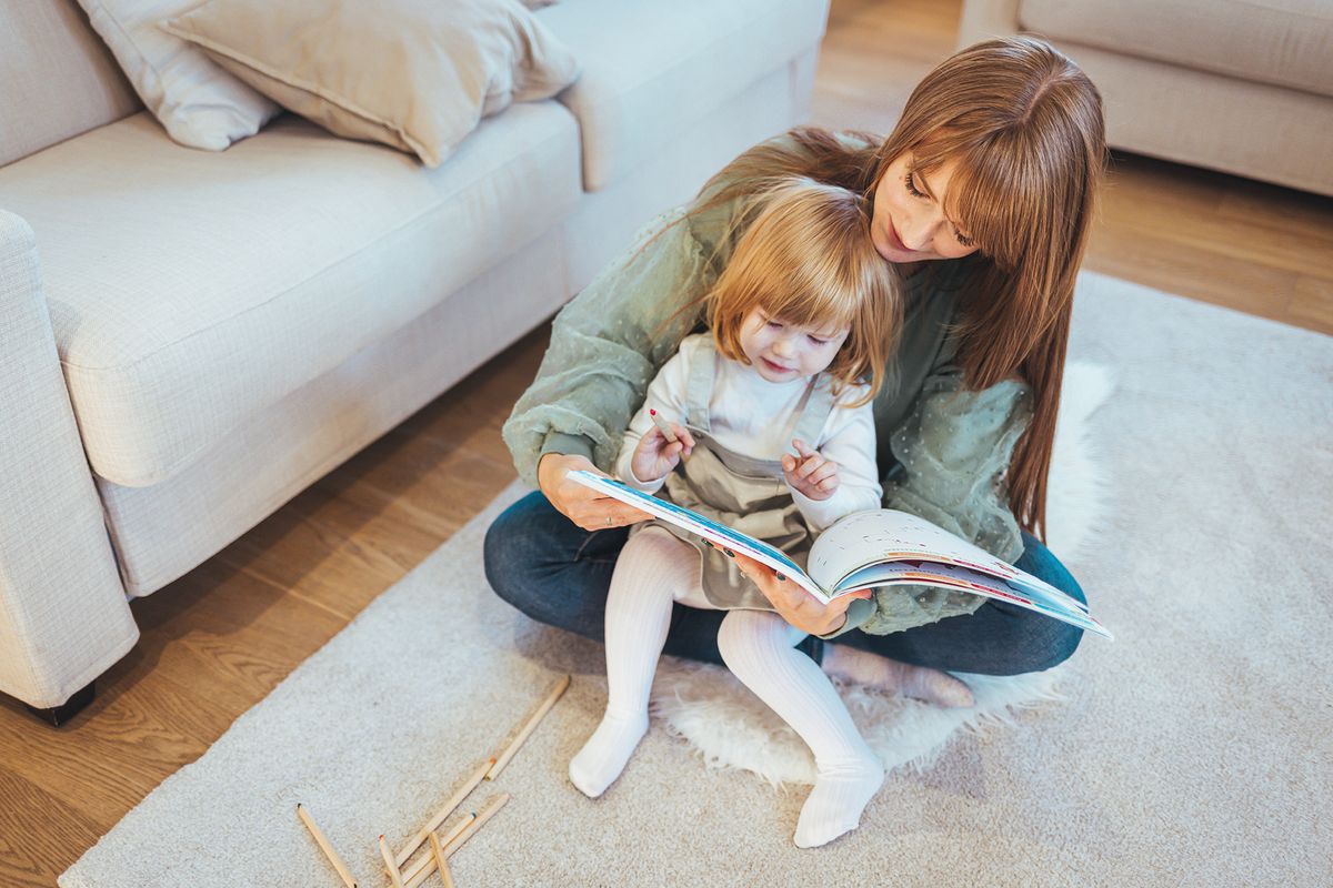 Portrait,Of,A,Smiling,Young,Cute,Mother,And,Daughter,ReadingPortrait of a smiling young cute mother and daughter reading a book lying and relax in the big white room. Mother teaching small daughter to drawing reading and writing