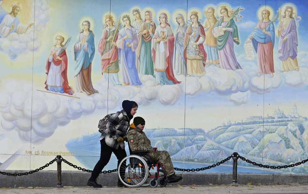 A girl carries a wheelchair with a disabled serviceman in front of frescoes of St. Michael's Golden-Domed Cathedral in Kyiv on November 20, 2023, amid the Russian invasion of Ukraine. (Photo by Sergei SUPINSKY / AFP)