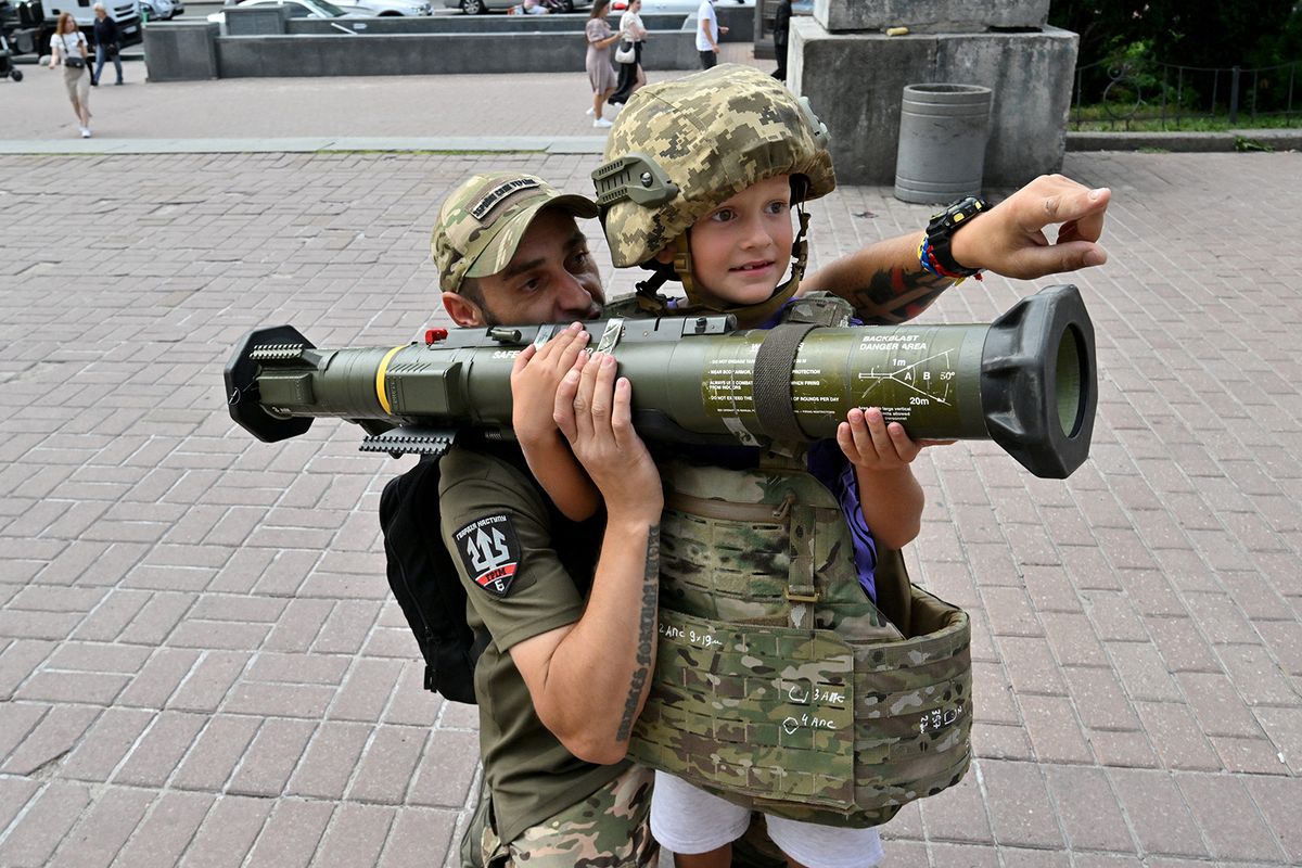 A Ukrainian serviceman helps a boy wearing body armour to hold and aim an AT4 Swedish man-portable anti-tank weapon at a volunteers point in the centre of Kyiv on August 10, 2023, amid Russian invasion in Ukraine. (Photo by Sergei SUPINSKY / AFP)