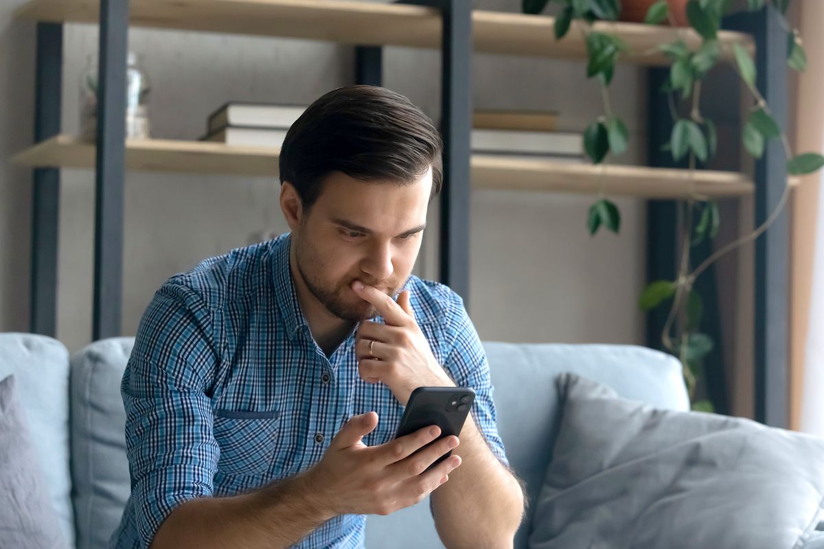 Thoughtful,Serious,Man,Touching,Chin,,Looking,At,Smartphone,Screen,,Sitting